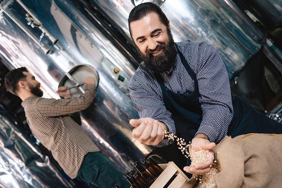 Specialized Business Insurance - Happy Bearded Man in Apron Pours Barley into his Hand While Another Man is Checking on a Tank in Their Distillery